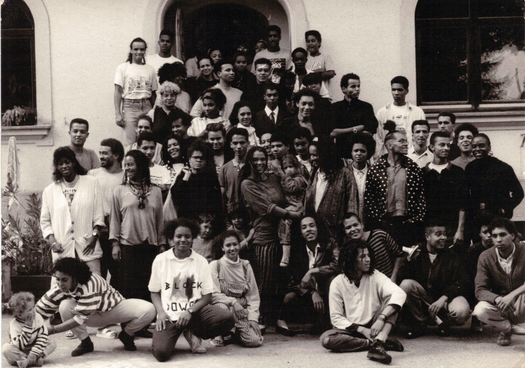Große Gruppe von Menschen unterschiedlichen Alters und Geschlechts vor einem Gebäude mit Bogenfenster, einige sitzen, andere stehen