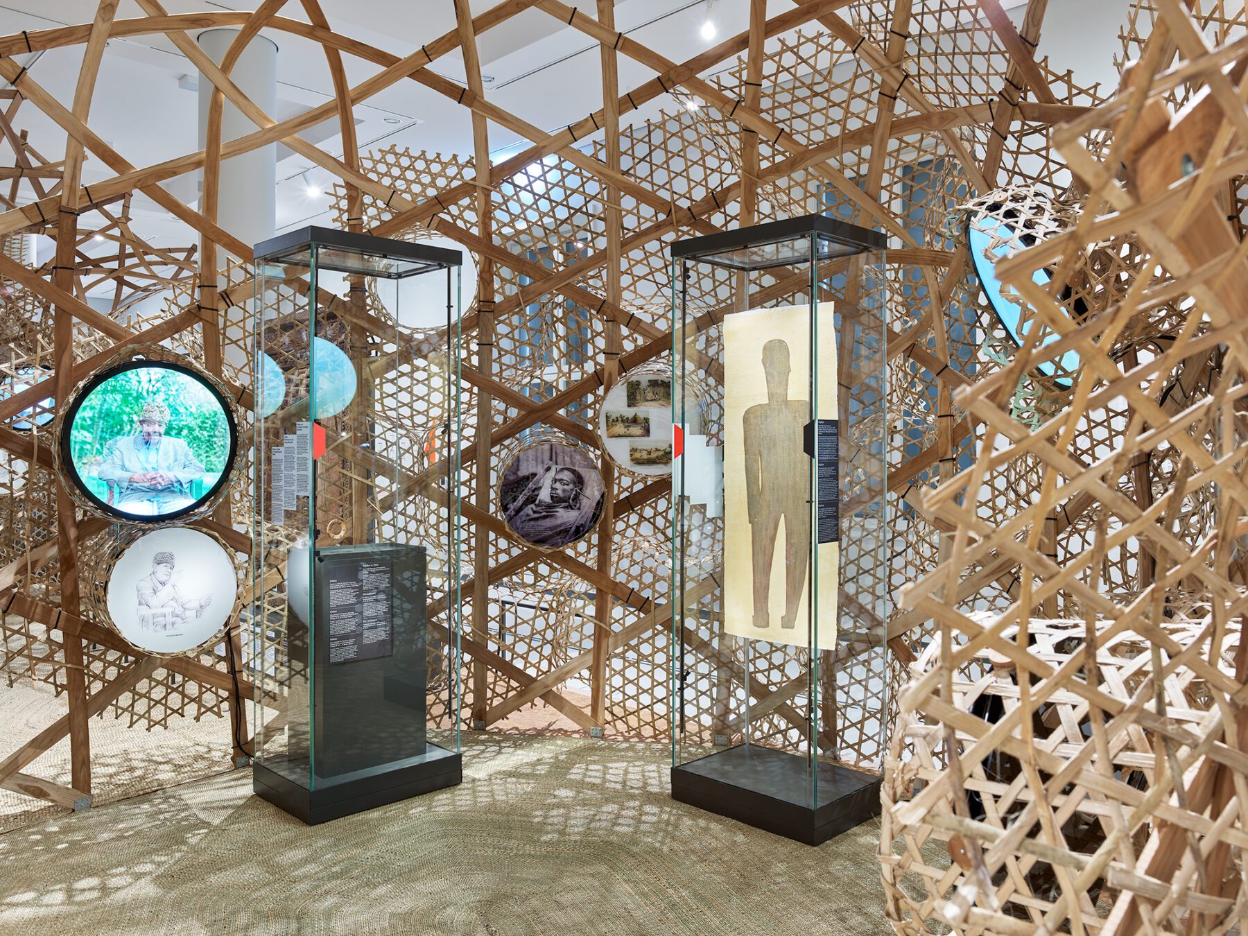 Ausstellungsraum mit zwei gläsernen Vitrinen, die jeweils eine Skulptur und Informationstafeln enthalten, umgeben von einem Gitter aus Holzlatten und mehreren runden Bildtafeln an den Wänden