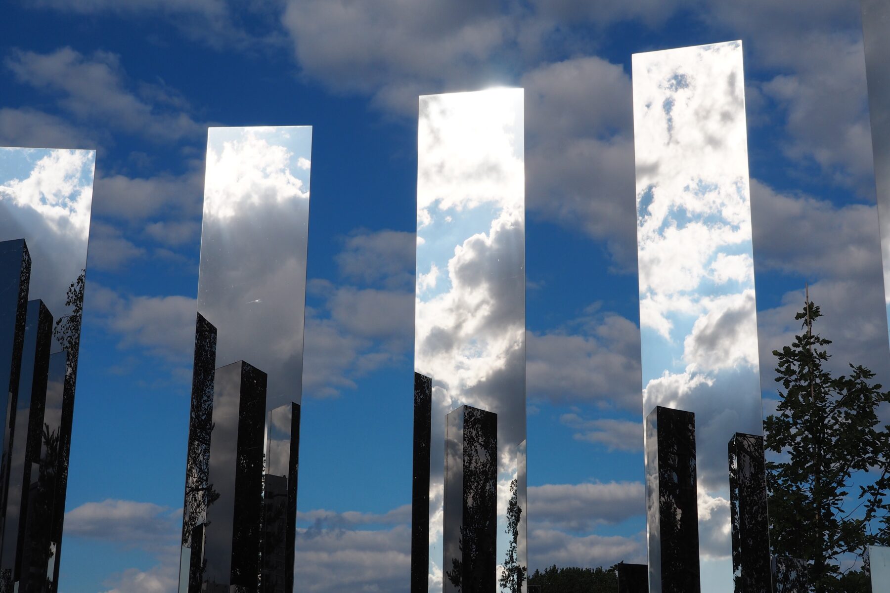 Mehrere hohe, schmale Spiegelstelen reflektieren den bewölkten Himmel vor einer Baumgruppe