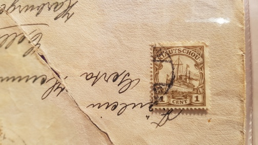 Nahaufnahme eines alten Briefumschlags mit einer braunen Briefmarke der Deutsch-Ostafrika-Kolonie mit dem Wert '1 Cent' und handschriftlichem Text im Hintergrund.