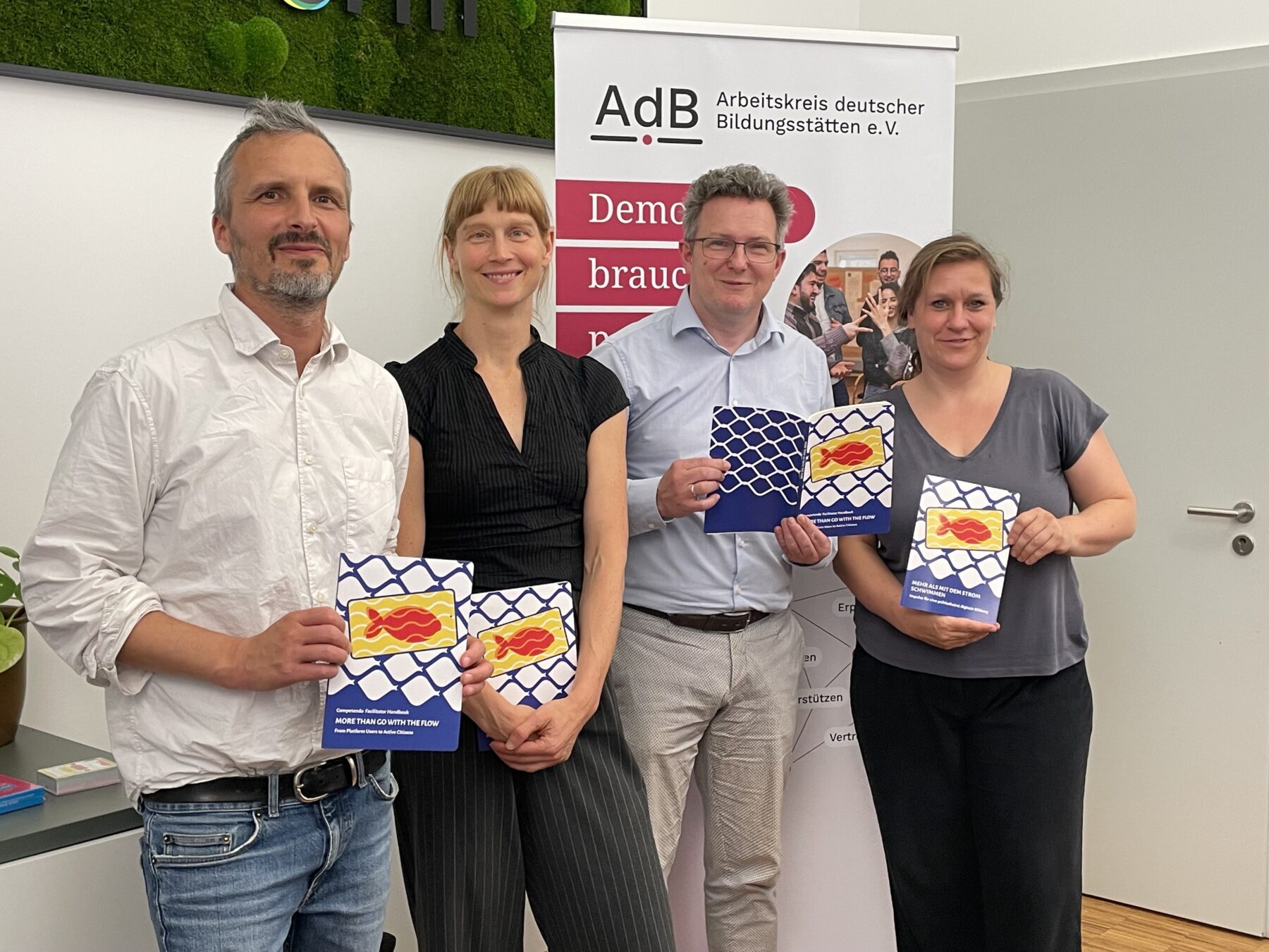Vier Personen stehen nebeneinander und halten jeweils ein blaues Heft mit einem roten Fisch auf weißem Netzmuster vor einem Banner des Arbeitskreises deutscher Bildungsstätten
