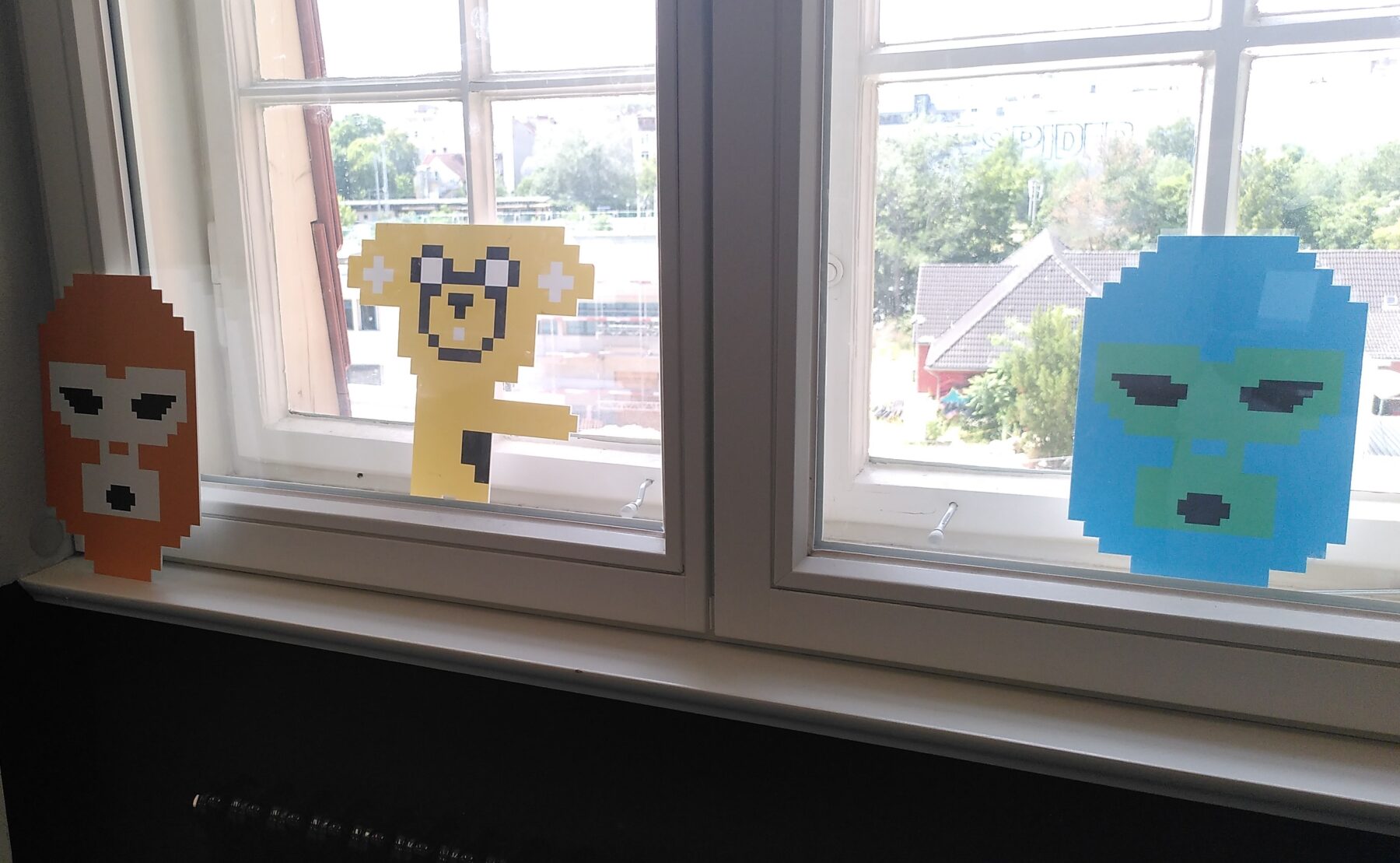 Drei bunte, pixelige Gesichter aus Papier auf einem Fensterbrett
