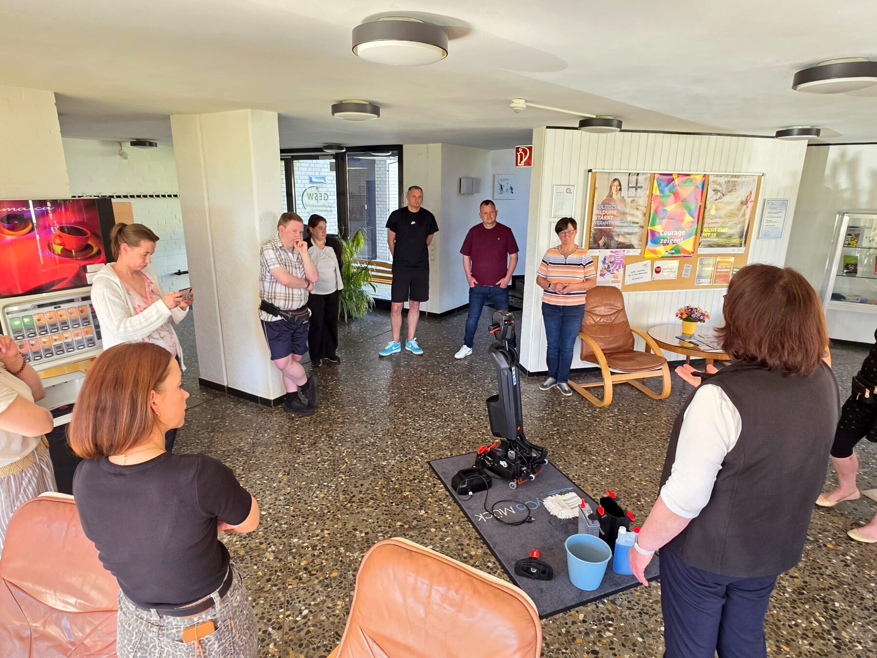 Gruppe von Menschen steht in einem Raum mit braunen Ledersesseln und mehreren bunten Postern an der Wand, eine Person spricht vor der Gruppe.